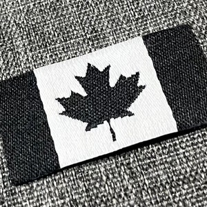 May include: Black and white Canadian flag patch with a maple leaf design. The patch is made of fabric and has a textured surface.