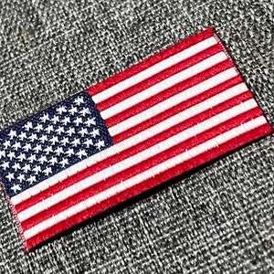 Könnte beinhalten: Ein kleiner, gestickter amerikanischer Flaggen-Patch mit roten und weißen Streifen und blauen Sternen. Der Patch befindet sich auf einem grauen Stoffhintergrund.