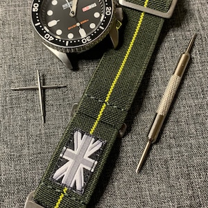 May include: A black and silver watch with a green and yellow striped NATO strap. The watch face has a black background with white markings and a date window. The strap has a Union Jack patch.