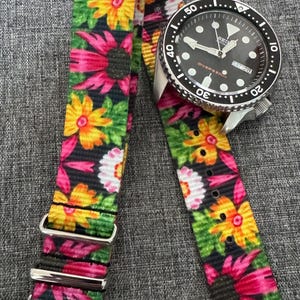 May include: A black wristwatch with a floral patterned watch strap. The strap displays a repeating pattern of pink, yellow, and white flowers on a black background. The watch face has a black bezel and shows the day and date.