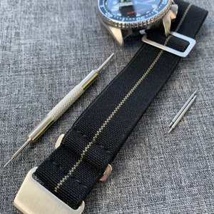 May include: A black and grey striped nylon watch strap with silver hardware and spring bars. The strap is laid out on a grey fabric surface with a watch and a tool for changing watch straps.
