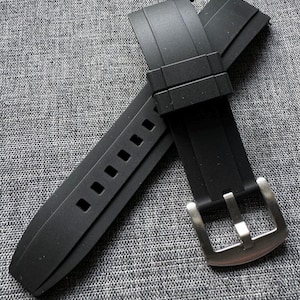 May include: A black rubber watch strap with a silver-colored buckle. The strap has a series of rectangular holes for adjustment. The buckle is rectangular with a single prong. The strap is lying on a gray textured surface.