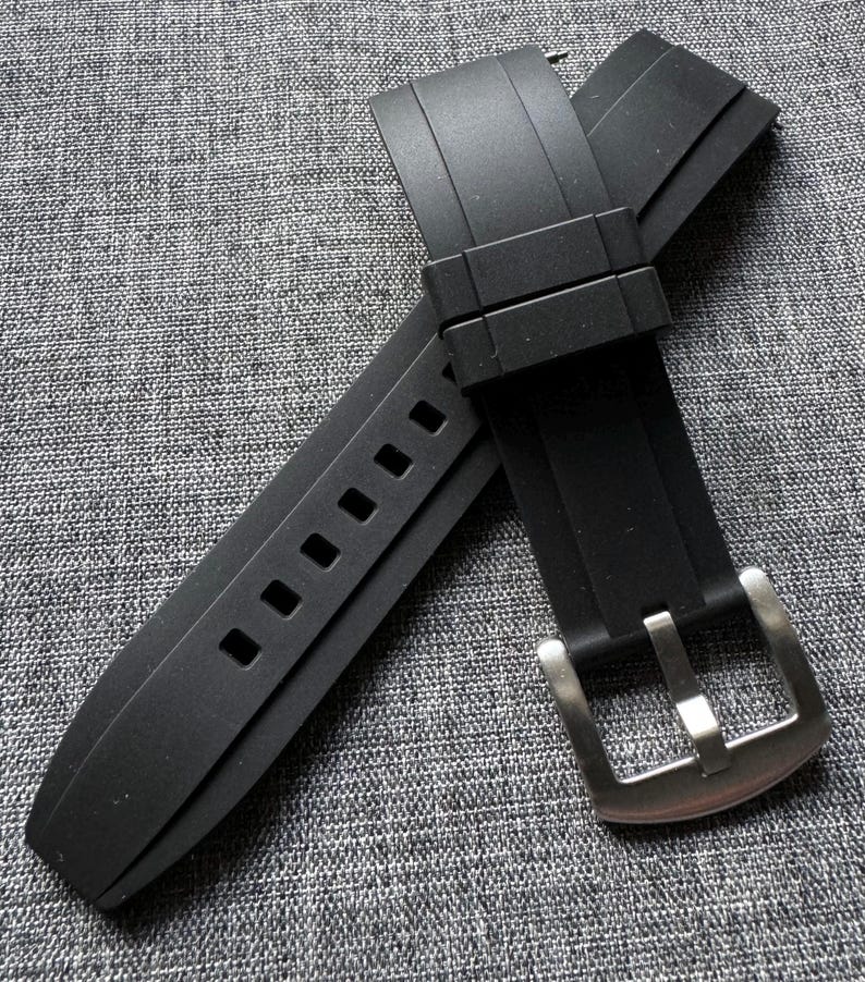 May include: A black rubber watch strap with a silver-colored buckle. The strap has a series of rectangular holes for adjustment. The buckle is rectangular with a single prong. The strap is lying on a gray textured surface.