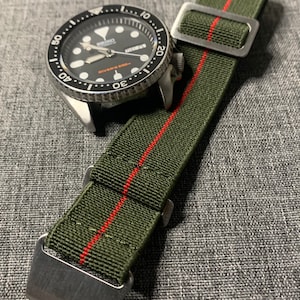 May include: A black and silver wristwatch with a green and red striped nylon watch strap. The watch face has white markings and the text "Divers 200m" printed on it.