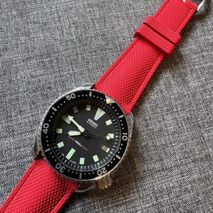 Puede incluir: Un reloj autom&aacute;tico Seiko con esfera negra y correa roja. El reloj tiene una caja de acero inoxidable, un bisel negro giratorio y agujas e indicadores luminosos. La correa roja es texturizada y tiene una hebilla plateada.