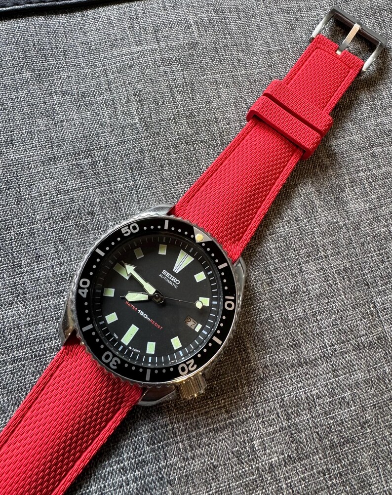 Puede incluir: Un reloj autom&aacute;tico Seiko con esfera negra y correa roja. El reloj tiene una caja de acero inoxidable, un bisel negro giratorio y agujas e indicadores luminosos. La correa roja es texturizada y tiene una hebilla plateada.