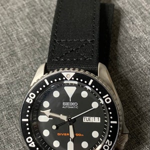 May include: A black Seiko automatic diver watch with a black canvas strap. The watch face has white markings and a date window that reads "TUE 11".