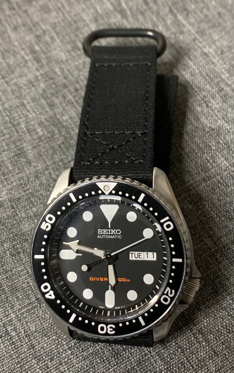 May include: A black Seiko automatic diver watch with a black canvas strap. The watch face has white markings and a date window that reads "TUE 11".