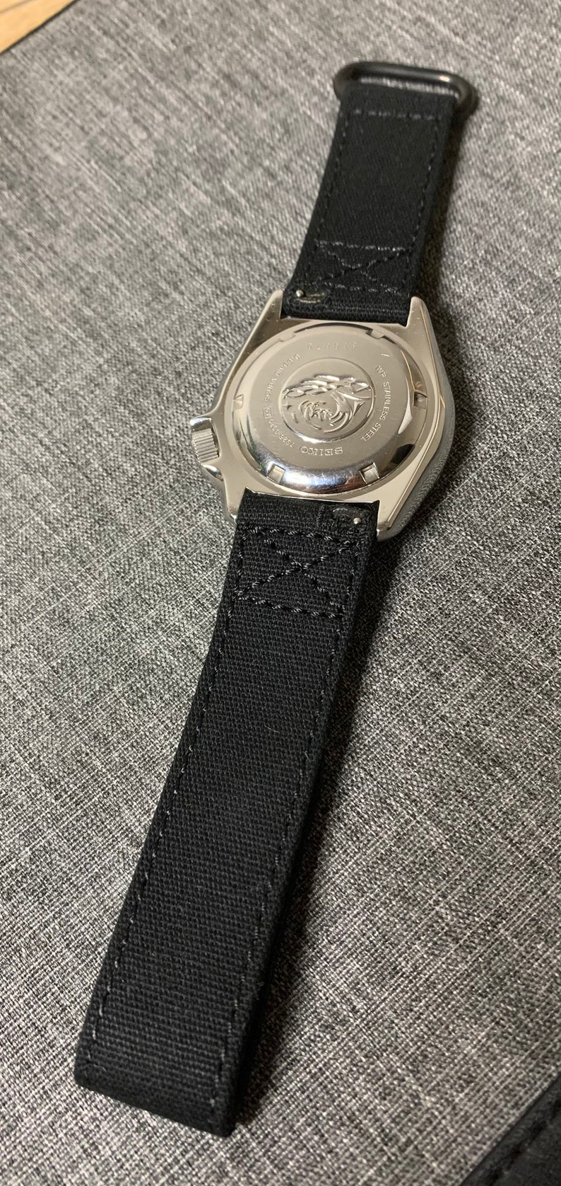 May include: A silver watch with a black fabric strap. The watch face is not visible. The back of the watch has the text "SEIKO" and "WATER RESIST" printed on it.