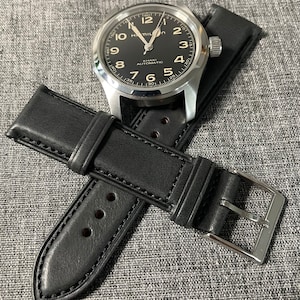 Puede incluir: Una correa de reloj de cuero negro con una hebilla plateada. La correa está unida a un reloj plateado con una esfera negra y agujas plateadas. La esfera del reloj tiene las palabras "Hamilton Khaki Automatic" impresas.