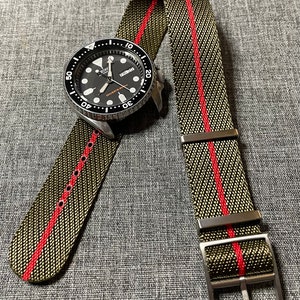 Könnte beinhalten: Eine schwarze und silberne Uhr mit schwarzem Zifferblatt und weißen Markierungen. Die Uhr ist an einem grün-rot gestreiften Nylon-Uhrenarmband.