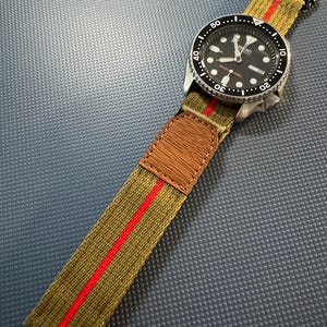 Puede incluir: Un reloj de pulsera negro y plateado con una esfera negra y una correa de nylon a rayas verdes y rojas.