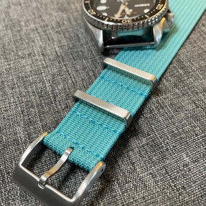 May include: A light blue nylon watch strap with silver buckle and keeper loops. The strap is made of a woven fabric with a subtle texture.
