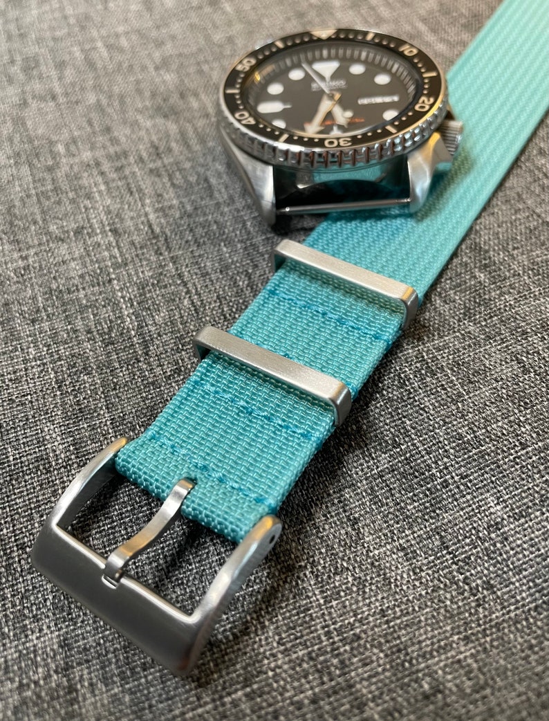 May include: A light blue nylon watch strap with silver buckle and keeper loops. The strap is made of a woven fabric with a subtle texture.