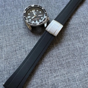 Puede incluir: Una correa de reloj de goma negra con un cierre plateado junto a un reloj plateado y negro. La esfera del reloj es negra con marcas y agujas blancas. La correa del reloj est&aacute; en &aacute;ngulo sobre una superficie gris texturizada.