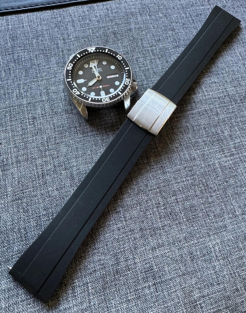 Puede incluir: Una correa de reloj de goma negra con un cierre plateado junto a un reloj plateado y negro. La esfera del reloj es negra con marcas y agujas blancas. La correa del reloj est&aacute; en &aacute;ngulo sobre una superficie gris texturizada.