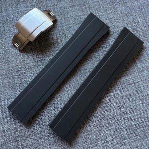 Puede incluir: Un cierre de reloj de acero inoxidable y dos correas de reloj negras se muestran sobre una superficie gris texturizada. El cierre del reloj es rectangular con un acabado de metal cepillado. Las correas del reloj son largas y rectangulares.