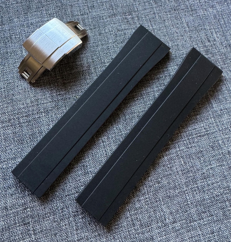 Puede incluir: Un cierre de reloj de acero inoxidable y dos correas de reloj negras se muestran sobre una superficie gris texturizada. El cierre del reloj es rectangular con un acabado de metal cepillado. Las correas del reloj son largas y rectangulares.