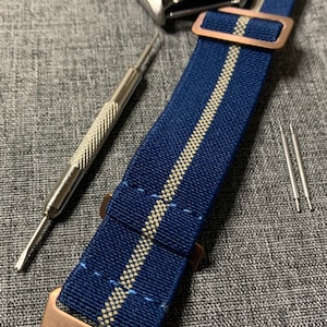 Op de afbeelding: Een blauw en wit gestreept nylon horlogebandje met een roségouden gesp. Het bandje ligt op een grijs stoffen oppervlak met twee kleine zilveren gereedschappen.