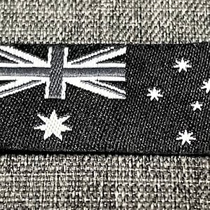 Könnte beinhalten: Ein schwarzer Stoff-Patch mit einer weißen gestickten australischen Flagge. Die Flagge zeigt das Union Jack in der oberen linken Ecke und eine Konstellation von sieben weißen Sternen in der unteren rechten Ecke.