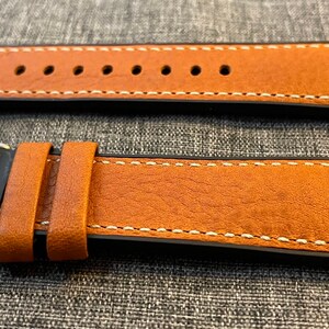 May include: A brown leather watch strap with white stitching and a black buckle. The strap is shown in two positions, one with the buckle attached and one with the buckle detached.
