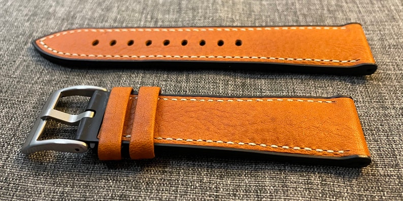 May include: A brown leather watch strap with white stitching and a black buckle. The strap is shown in two positions, one with the buckle attached and one with the buckle detached.