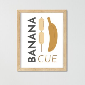May include: A framed print with the text "BANANA CUE" in black and a graphic of a banana and a skewer with three small, round, beige items on it.