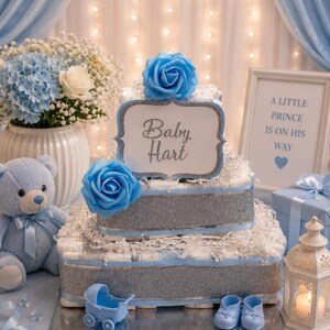 May include: A two-tier nappy cake decorated with silver glitter ribbon, blue roses, and a sign reading Baby Hart. The display includes a light blue teddy bear, a miniature blue baby carriage, a pair of blue booties, and a framed sign stating A LITTLE PRINCE IS ON HIS WAY.