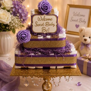 May include: A two-tiered nappy cake featuring white nappies, gold glitter ribbons, and purple satin ribbons. It is decorated with purple foam roses and purple paper shred. A sign reads Welcome Sweet Baby Bri! It sits on a gold pedestal cake stand.