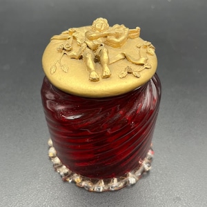 Può includere: Un vaso decorativo con un corpo in vetro rosso a spirale e un coperchio color oro. Il coperchio presenta una scultura a rilievo di un cherubino che suona uno strumento a corde. La base del vaso ha un bordo trasparente e strutturato.