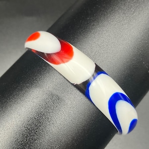 May include: A smooth, rounded bangle bracelet featuring a glossy white base with vibrant, swirling patterns of red and blue glass accents. The design consists of opaque white segments interspersed with bold, colorful patches of red and blue.