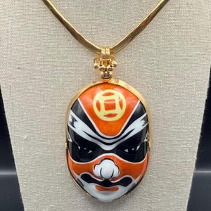 May include: A gold-toned necklace featuring a pendant shaped like a Japanese Noh mask. The mask is painted with orange, black, white, and yellow details. The pendant is suspended from a gold chain, creating a striking piece of jewelry.