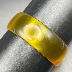 May include: A smooth, translucent yellow resin bangle bracelet with a polished finish. The band features a consistent, rounded rectangular profile with a vibrant, honey-colored hue that allows light to pass through the material, highlighting its clarity.