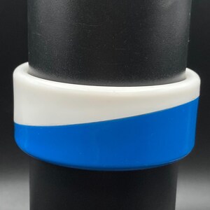 May include: A wide bangle bracelet featuring a smooth, glossy finish with a two-tone design. The upper portion is solid white, while the lower portion is a vibrant, solid blue. The two colors meet in a diagonal, curved wave pattern across the surface.