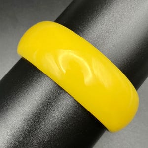 May include: A solid, bright yellow bangle bracelet with a smooth, glossy finish. The bracelet has a rounded, thick profile and a uniform, vibrant colour throughout its entire surface, reflecting light across its curved, polished exterior.