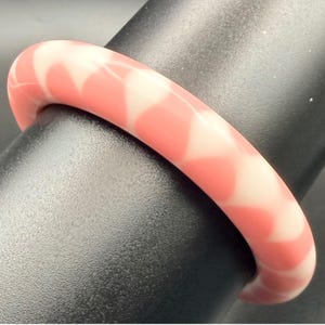 May include: A smooth, rounded bangle bracelet featuring a marbled pattern of soft coral pink and creamy white colors. The circular accessory has a polished, glossy finish and a continuous, solid band design with no visible clasps or openings.