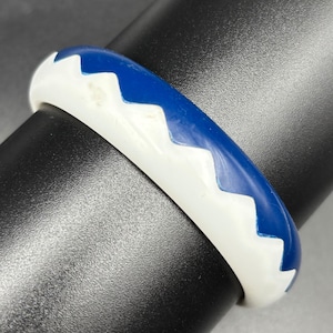 Puede incluir: Una pulsera rígida circular con un diseño bicolor que presenta una base blanca lisa y un patrón de zigzag en azul profundo que recorre toda la circunferencia de la pieza, creando un efecto visual geométrico muy definido.