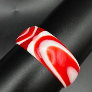 May include: A wide bangle bracelet featuring a smooth, glossy finish with a swirling, marbled pattern of vibrant red and opaque white colors. The bracelet has a rounded, thick profile and a continuous, seamless circular band design.
