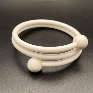 May include: A white spiral bracelet featuring a smooth, continuous band that wraps around to form two full loops. Each end of the bracelet is finished with a small, solid white spherical bead, creating a clean and uniform appearance throughout.