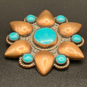 Peut inclure: Une broche circulaire en cuivre avec un grand cabochon turquoise central entouré de six pétales de cuivre en forme de goutte et de six cabochons turquoise ronds plus petits. Le métal présente des bordures décoratives torsadées.