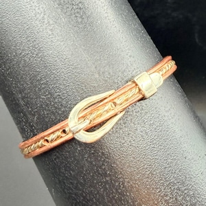 Puede incluir: Un anillo de cinturón de cuero marrón con una hebilla dorada. El anillo presenta un pequeño diseño de cinturón decorativo con una hebilla dorada y una banda trenzada dorada. El anillo está diseñado para usarse en un dedo.
