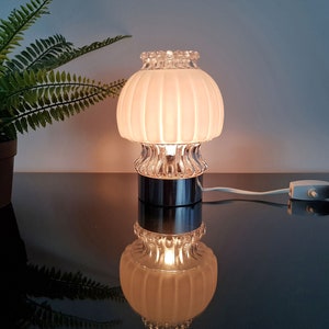 Könnte beinhalten: Eine weiße Tischlampe aus Glas mit einem gerippten Schirm und einem verchromten Fuß. Die Lampe ist eingeschaltet und leuchtet.