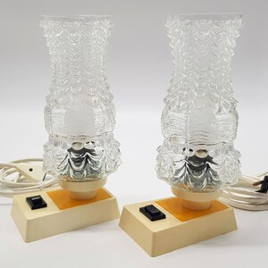 Könnte beinhalten: Zwei Vintage-Tischlampen mit klaren Glasschirmen mit einem strukturierten, gewellten Design. Jede Lampe hat einen weißen Sockel mit einem gelben Akzent und einem schwarzen Ein-/Ausschalter.