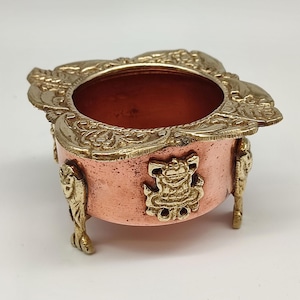 Puede incluir: Un pequeño cuenco adornado de color cobre con detalles dorados. El cuenco tiene un borde decorativo y está sostenido por cuatro pequeños pies dorados. Los lados del cuenco presentan adornos dorados. El cuenco está vacío y descansa sobre una superficie blanca.
