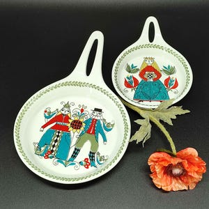 Könnte beinhalten: Zwei weiße Keramik-Servierschalen mit Griffen. Jede Schale hat ein farbenfrohes Volkskunstdesign mit Menschen und Blumen. Die Ränder sind mit einem grünen Blättermuster verziert. Neben einer Schale befindet sich eine rot-orangefarbene Blume.
