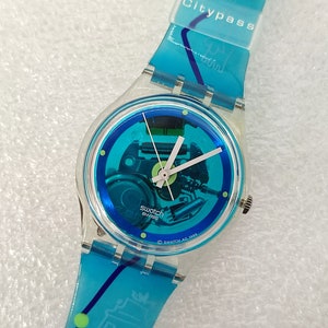 Könnte beinhalten: Eine blaue und silberne Swatch-Uhr mit einem durchsichtigen Plastikband. Das Band trägt den Text "Citypass". Das Zifferblatt der Uhr hat ein blaues und grünes Design mit einem sichtbaren Mechanismus.