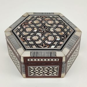 Könnte beinhalten: Eine achteckige Holzbox mit aufwendigen Einlegearbeiten aus Perlmutt und geometrischen Mustern. Die Box hat einen dunkelbraunen Sockel mit dekorativen Paneelen und einen kontrastierenden hellfarbigen Deckel mit Sternmuster. Die Kanten sind mit schwarz-weißen Streifen versehen.