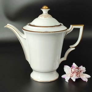Könnte beinhalten: Weiße Porzellan-Kaffeekanne mit goldener Verzierung und einem Blumendesign auf dem Deckel. Die Kanne hat einen runden Korpus und einen Ausguss an der Seite. Der Griff ist ebenfalls golden.
