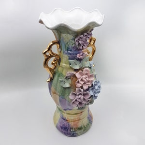 Könnte beinhalten: Eine Keramikvase mit einem weißen, gewellten oberen Rand und goldenen Griffen. Die Vase ist mit einem bunten Blumenmuster und 3D-Blumen in Rosa-, Blau- und Grüntönen verziert.