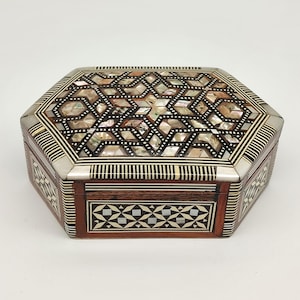Könnte beinhalten: Eine achteckige Holzbox mit aufwendigen Einlegearbeiten aus Perlmutt und geometrischen Mustern. Die Box hat einen dunklen Holzsockel, silberfarbene Metallverzierungen und einen dekorativen Deckel. Das Design umfasst Stern- und Diamantformen.